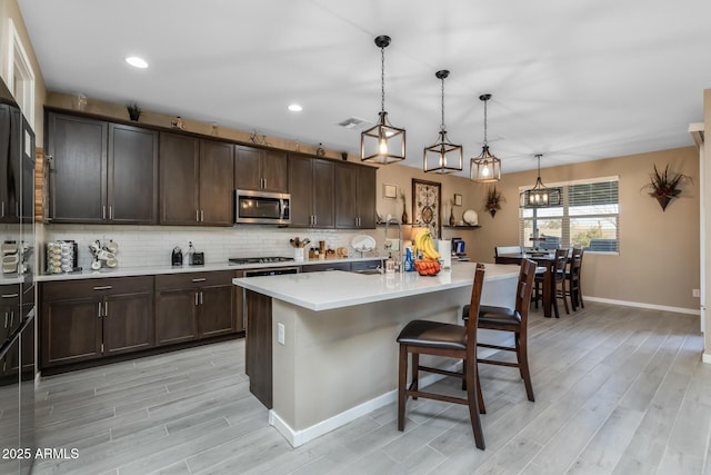 kitchen featuring a kitchen island with sink, hanging light fixtures, backsplash, dark brown cabinets, and stainless steel appliances