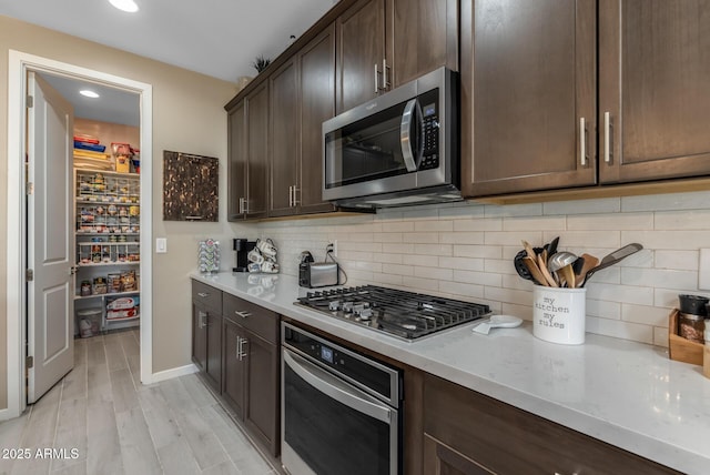 kitchen with appliances with stainless steel finishes, backsplash, light stone countertops, dark brown cabinets, and light wood-type flooring