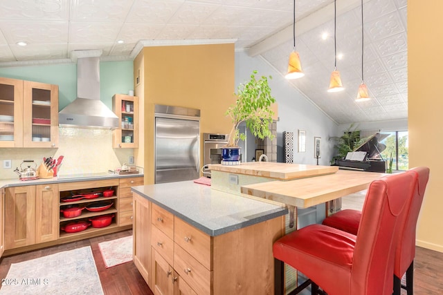 kitchen with wall chimney range hood, appliances with stainless steel finishes, hanging light fixtures, a kitchen bar, and light brown cabinets