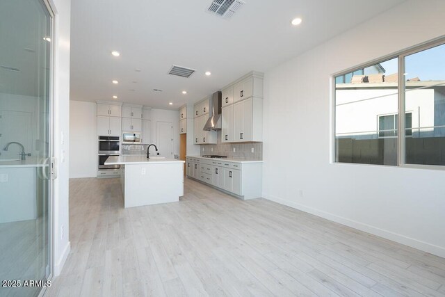 kitchen with wall chimney exhaust hood, an island with sink, decorative backsplash, black gas cooktop, and sink