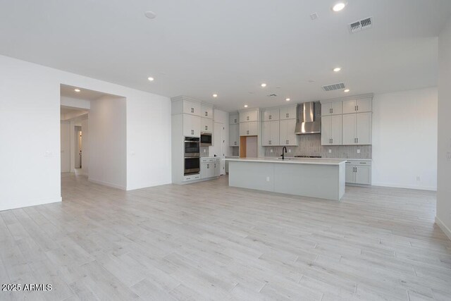 kitchen featuring a center island with sink, light hardwood / wood-style flooring, wall chimney range hood, stainless steel appliances, and tasteful backsplash