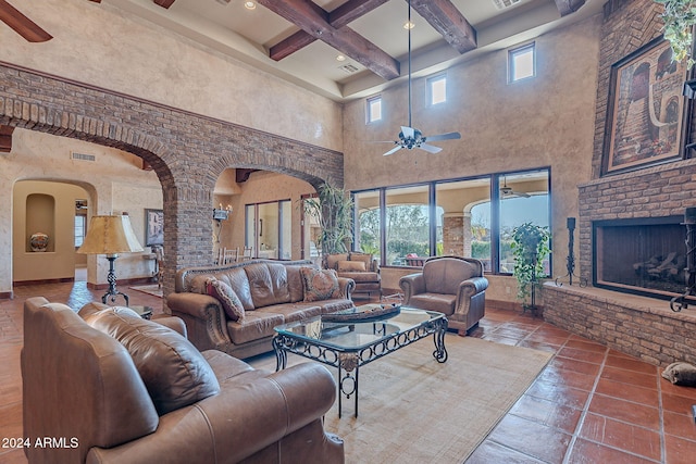 tiled living room featuring beam ceiling, ceiling fan, coffered ceiling, a high ceiling, and a brick fireplace