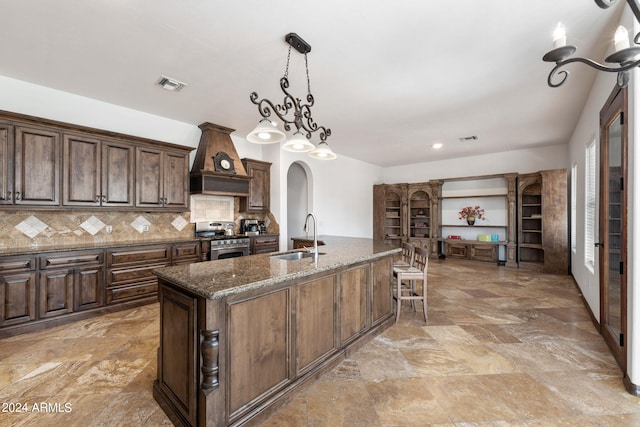 kitchen with stainless steel range with gas cooktop, a center island with sink, premium range hood, sink, and dark stone counters