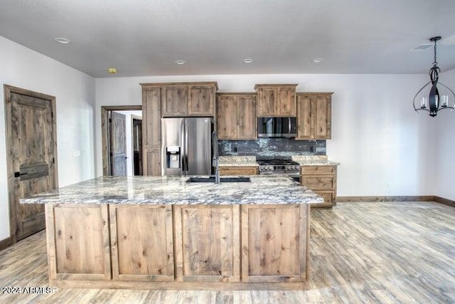 kitchen with light wood finished floors, decorative backsplash, appliances with stainless steel finishes, light stone countertops, and a kitchen island with sink