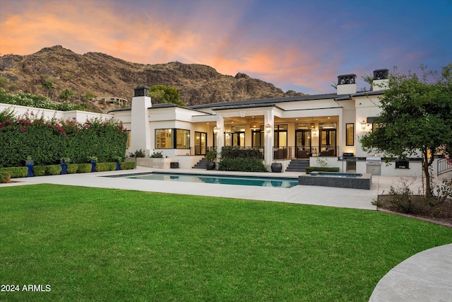 back house at dusk featuring a mountain view, a swimming pool with hot tub, a lawn, and a patio area