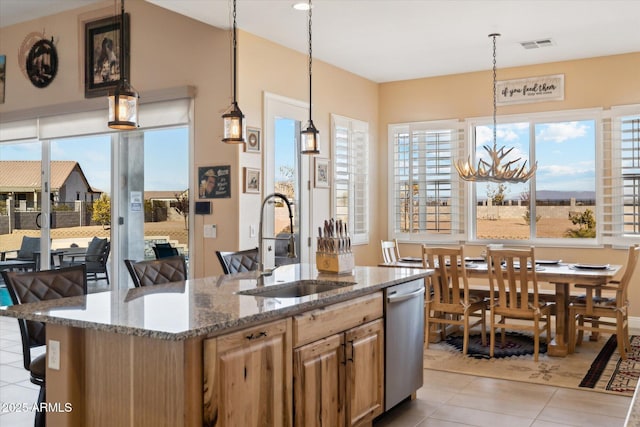 kitchen with an island with sink, sink, hanging light fixtures, light stone counters, and stainless steel dishwasher