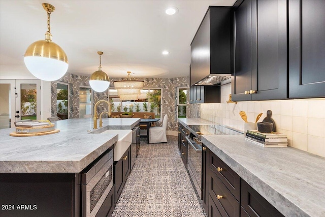 kitchen with wall chimney range hood, tasteful backsplash, a center island with sink, hanging light fixtures, and sink