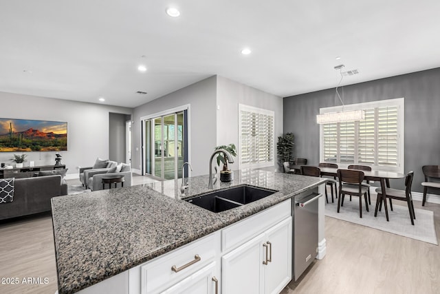 kitchen with a sink, light wood-type flooring, dark stone countertops, stainless steel dishwasher, and a kitchen island with sink