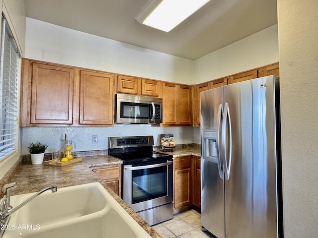 kitchen with stainless steel appliances, light tile patterned floors, brown cabinetry, and a sink