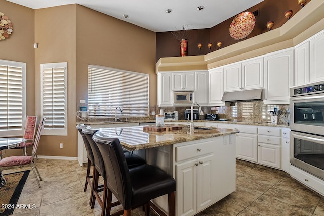 kitchen with appliances with stainless steel finishes, a towering ceiling, light stone counters, white cabinets, and an island with sink