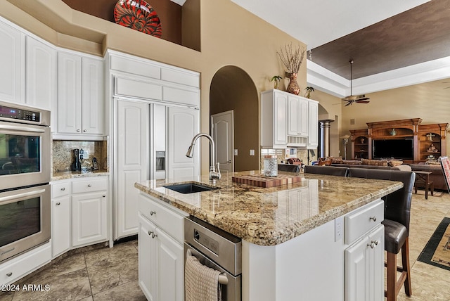 kitchen featuring sink, double oven, a towering ceiling, a kitchen island with sink, and white cabinets