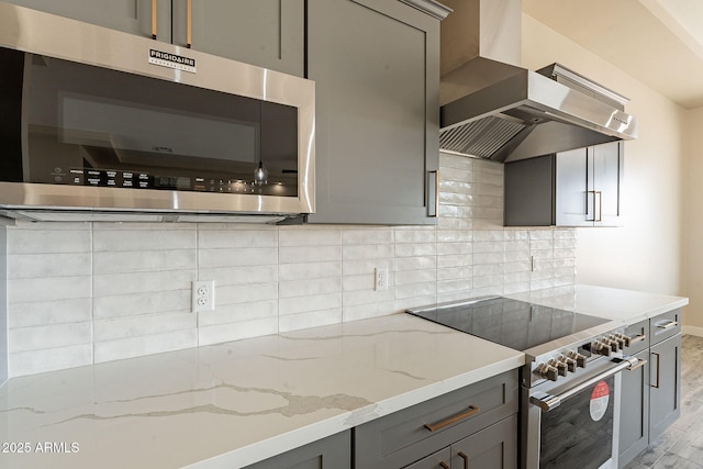 kitchen featuring light wood finished floors, wall chimney range hood, light stone countertops, gray cabinetry, and appliances with stainless steel finishes
