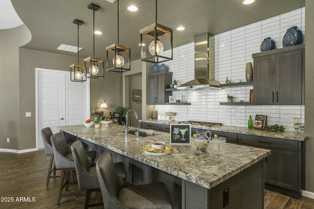 kitchen featuring light stone countertops, sink, hanging light fixtures, wall chimney range hood, and a kitchen island with sink