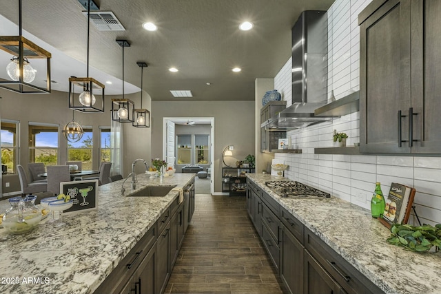 kitchen featuring stainless steel gas stovetop, wall chimney range hood, sink, light stone countertops, and a notable chandelier