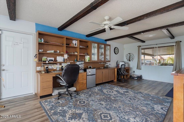 office with ceiling fan, built in desk, a textured ceiling, beamed ceiling, and light wood-type flooring