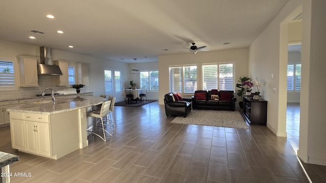 interior space featuring open floor plan, wall chimney exhaust hood, a center island with sink, and pendant lighting