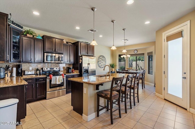 kitchen with stainless steel appliances, light stone countertops, an island with sink, glass insert cabinets, and pendant lighting