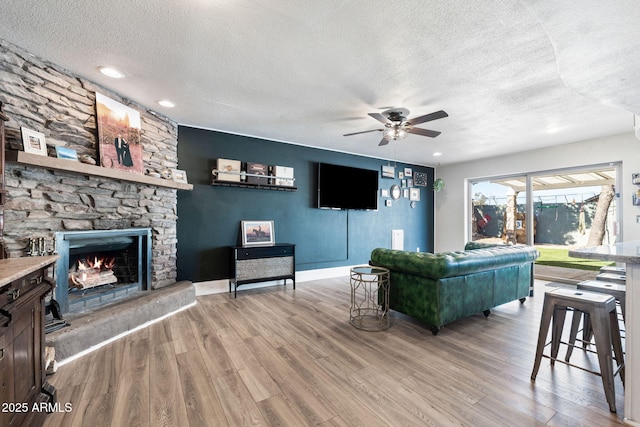 living room featuring ceiling fan, a stone fireplace, a textured ceiling, and light wood-type flooring