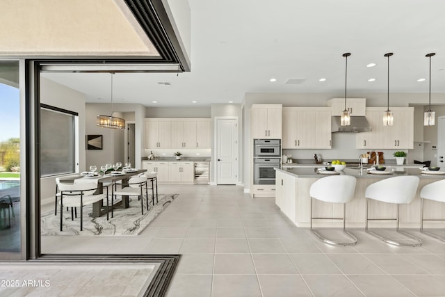 kitchen with double oven, light tile patterned floors, decorative light fixtures, a center island with sink, and a notable chandelier