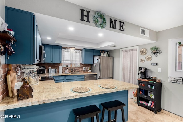 kitchen with blue cabinets, appliances with stainless steel finishes, a tray ceiling, kitchen peninsula, and a kitchen breakfast bar