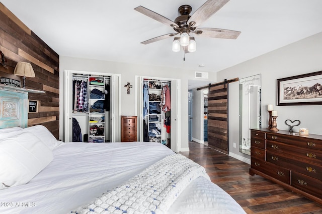 bedroom featuring ceiling fan, wooden walls, dark hardwood / wood-style flooring, two closets, and a barn door