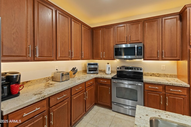 kitchen with stainless steel appliances, brown cabinets, light stone counters, and light tile patterned floors