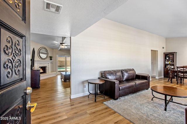 living room with a warm lit fireplace, light wood-style flooring, visible vents, and a ceiling fan