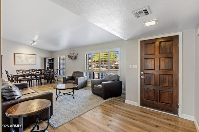 living area featuring baseboards, visible vents, vaulted ceiling, a textured ceiling, and light wood-type flooring