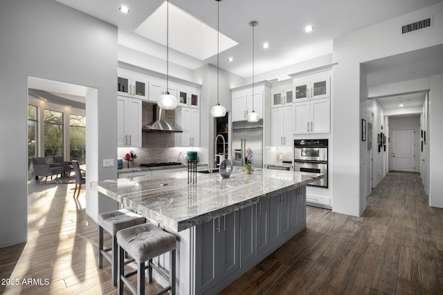 kitchen with dark wood-style floors, visible vents, appliances with stainless steel finishes, wall chimney range hood, and a large island with sink