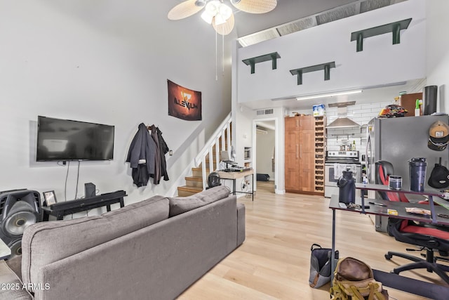 living area with ceiling fan, a high ceiling, visible vents, light wood-style floors, and stairs