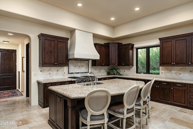 kitchen featuring dark brown cabinets, a center island with sink, light stone countertops, and premium range hood