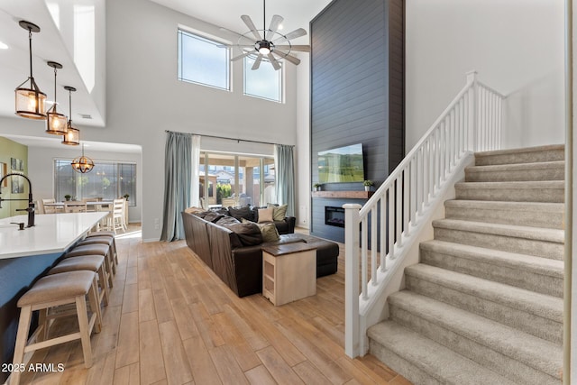 living room with sink, light hardwood / wood-style flooring, a large fireplace, ceiling fan with notable chandelier, and a high ceiling