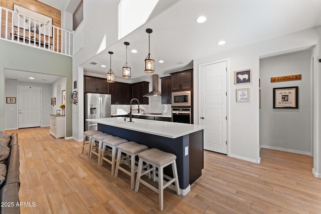 kitchen featuring pendant lighting, dark brown cabinets, stainless steel appliances, an island with sink, and wall chimney exhaust hood