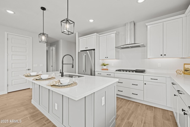 kitchen with stainless steel appliances, wall chimney range hood, light wood-style flooring, a center island with sink, and recessed lighting