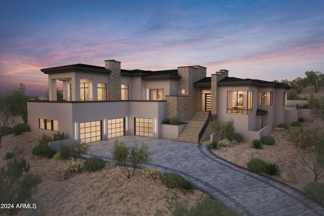 back of property at dusk with a garage, stone siding, a chimney, decorative driveway, and stucco siding