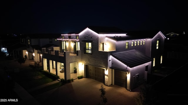 back of house at night with a standing seam roof, driveway, a balcony, an attached garage, and stone siding