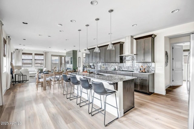 kitchen featuring a center island with sink, backsplash, decorative light fixtures, light stone countertops, and wall chimney exhaust hood