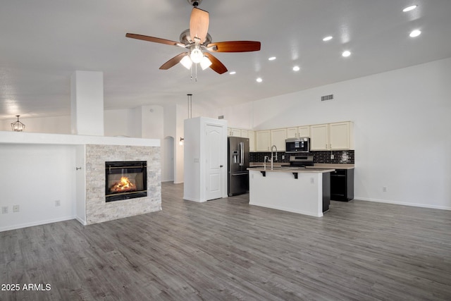 kitchen featuring a kitchen breakfast bar, a stone fireplace, a center island with sink, stainless steel appliances, and ceiling fan