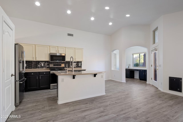 kitchen with a high ceiling, appliances with stainless steel finishes, an island with sink, sink, and light hardwood / wood-style flooring