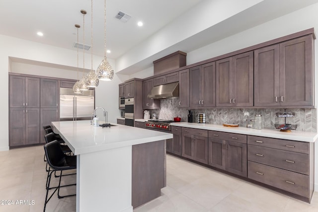 kitchen featuring appliances with stainless steel finishes, backsplash, a kitchen island with sink, sink, and decorative light fixtures