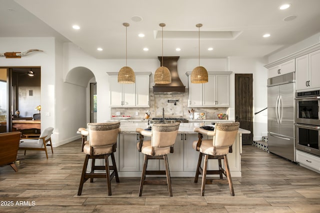 kitchen with decorative backsplash, arched walkways, wood finished floors, custom exhaust hood, and stainless steel appliances