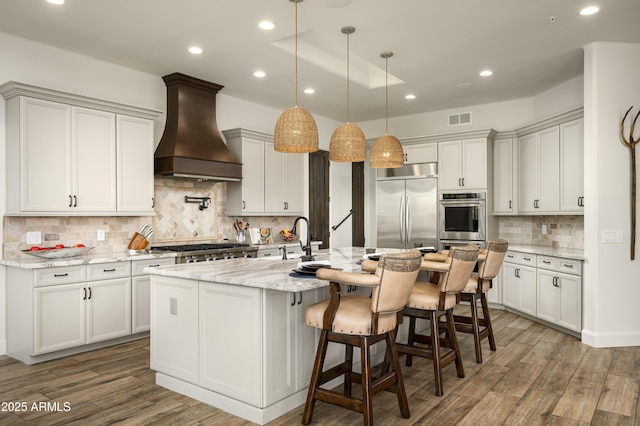 kitchen with wood finished floors, visible vents, premium range hood, an island with sink, and stainless steel appliances