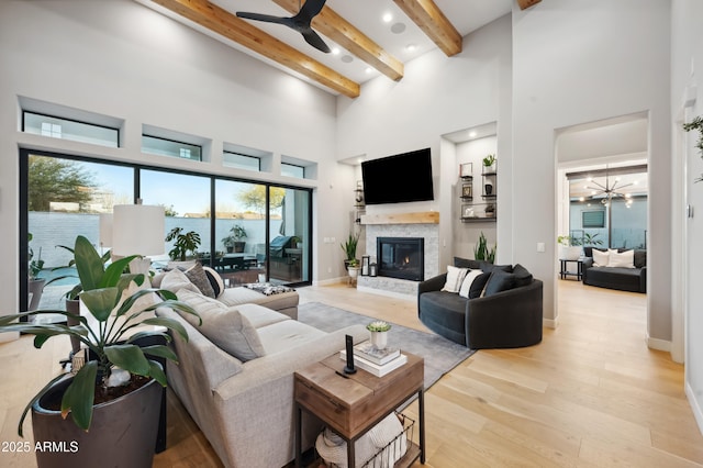 living room featuring beam ceiling, light wood-type flooring, a towering ceiling, a fireplace, and ceiling fan with notable chandelier