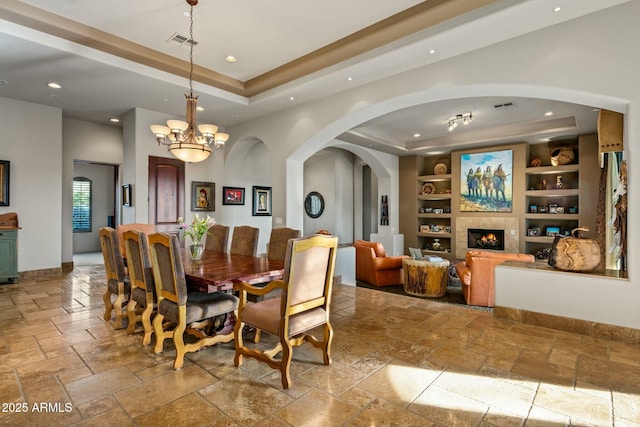 dining room featuring built in features, a tray ceiling, stone tile flooring, and a lit fireplace