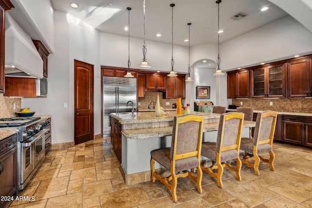 kitchen with stone tile flooring, a large island with sink, high quality appliances, and wall chimney exhaust hood