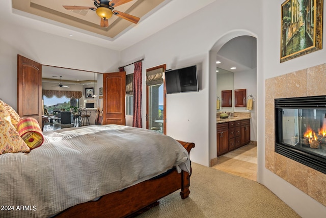 bedroom featuring connected bathroom, a tiled fireplace, light carpet, arched walkways, and a raised ceiling