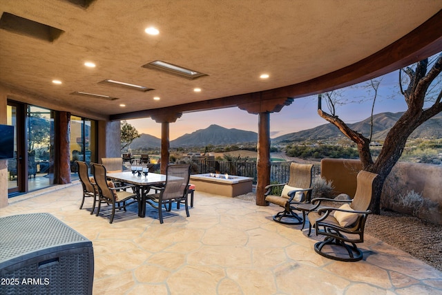 patio terrace at dusk featuring outdoor dining space, a mountain view, an outdoor fire pit, and fence