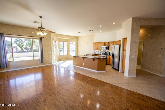 kitchen featuring light hardwood / wood-style floors, an island with sink, ceiling fan, appliances with stainless steel finishes, and a breakfast bar