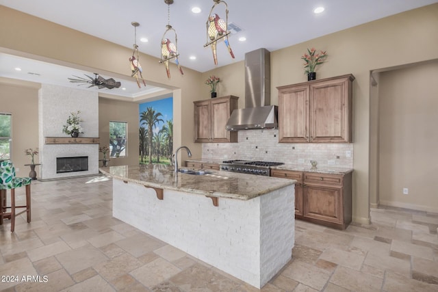 kitchen featuring a center island with sink, sink, wall chimney exhaust hood, ceiling fan, and light stone counters