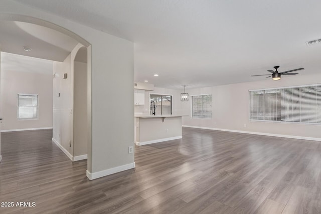 unfurnished living room with arched walkways, ceiling fan, visible vents, baseboards, and dark wood-style floors